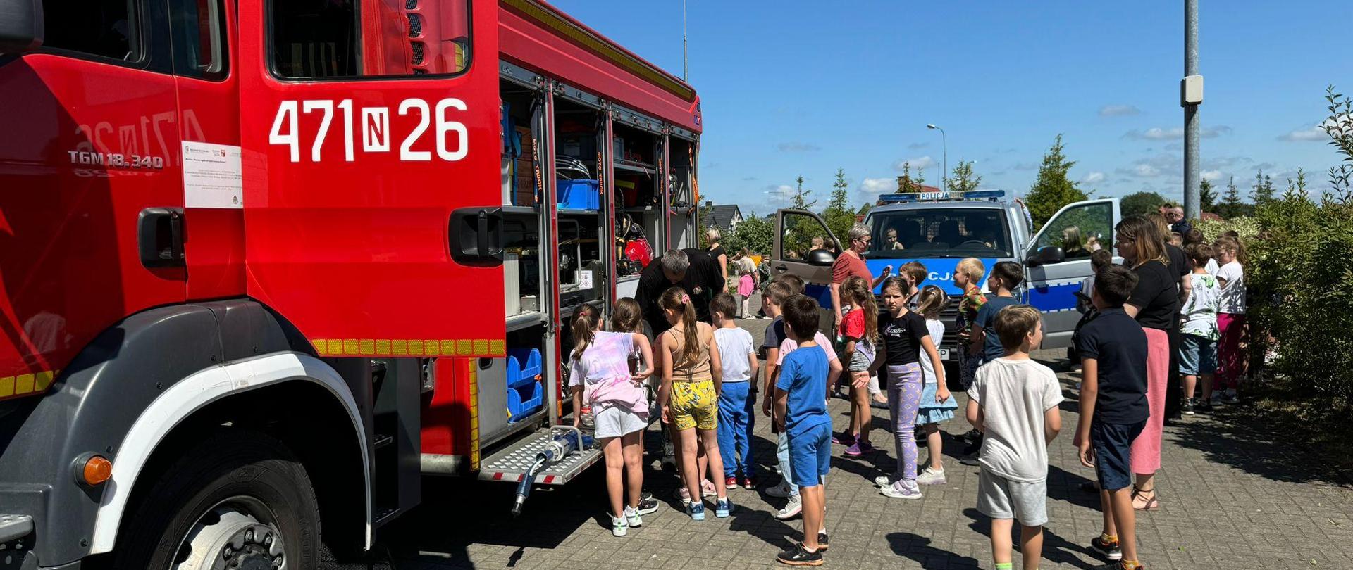 Na zdjęciu widać czerwony samochód strażacki, gromadkę dzieci i nauczyciela. Widać również pojazd Policji. 