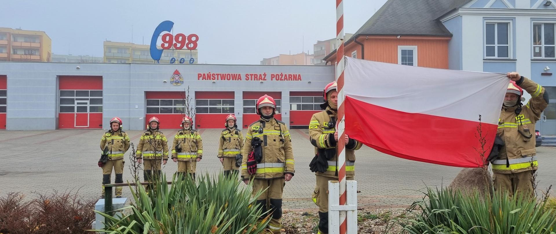 Strażacy stoją przed masztem flagowym na placu KP PSP Kwidzyn, zawieszają i podnoszą flagę