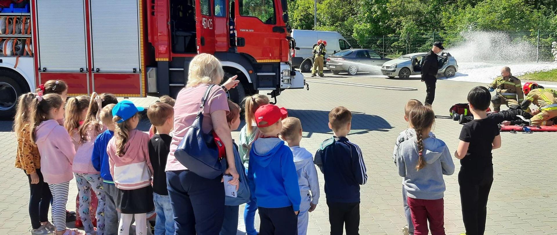 Zdjęcie przedstawia dzieci obserwujące pokaz ratownictwa drogowego wykonywany przez strażaków JRG w Pińczowie. 