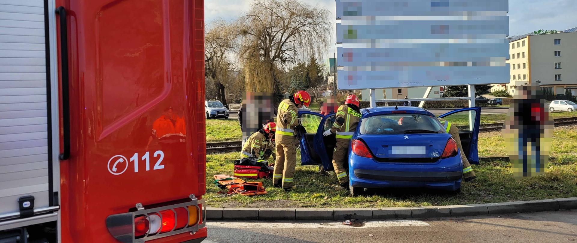 Na pierwszym planie strażacy podczas czynności ratowniczych, w tle uszkodzony pojazd i karetka pogotowia oraz wóz strażacki