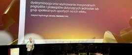 Wystąpienie Macieja Kmity z Departamentu Polityki Senioralnej KPRM - obok mównicy stoi mężczyzna w jasnej marynarce i ciemnych spodniach, w tle duży ekran z napisem "ageizm" i definicją tego pojęcia