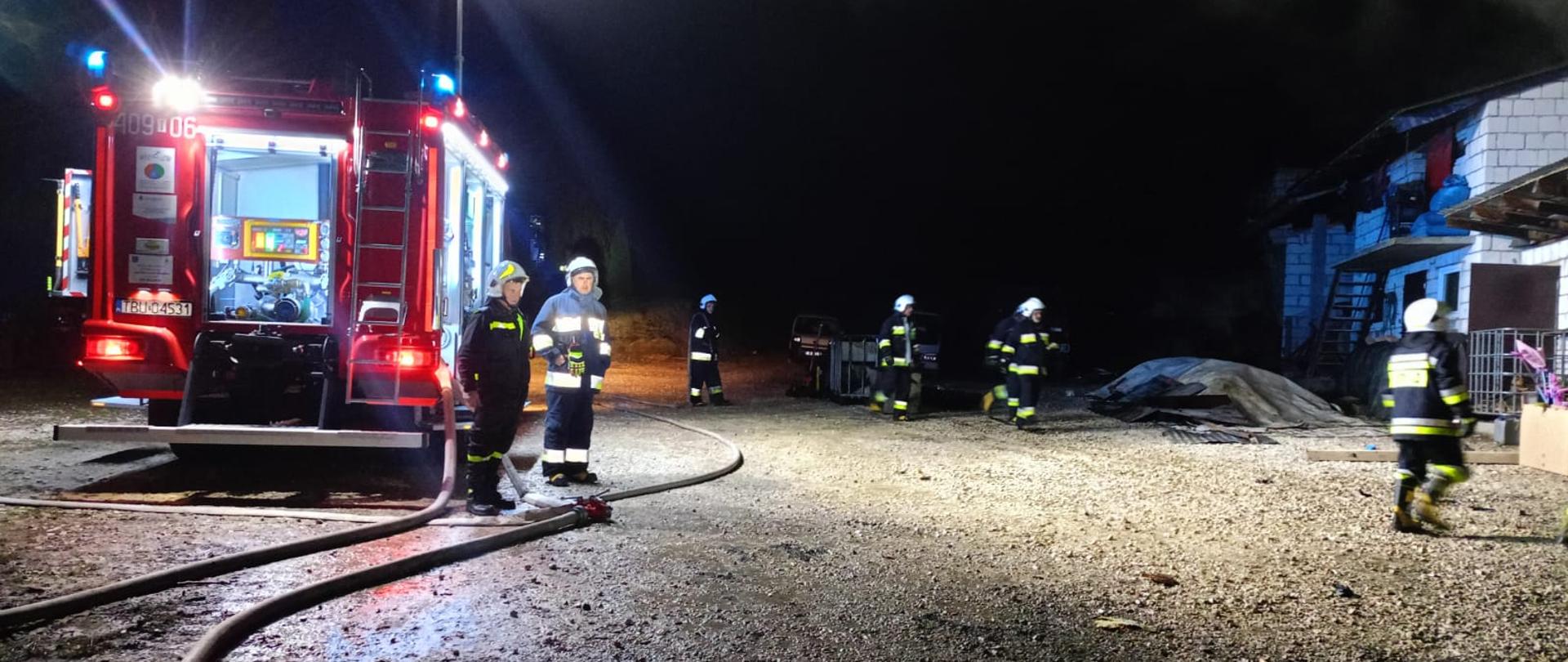 Po lewej stronie stoi samochód pożarniczy i od niego rozwinięte węże, przy samochodzie strażacy, po prawej stronie w tle strażacy i budynek gospodarczy. 