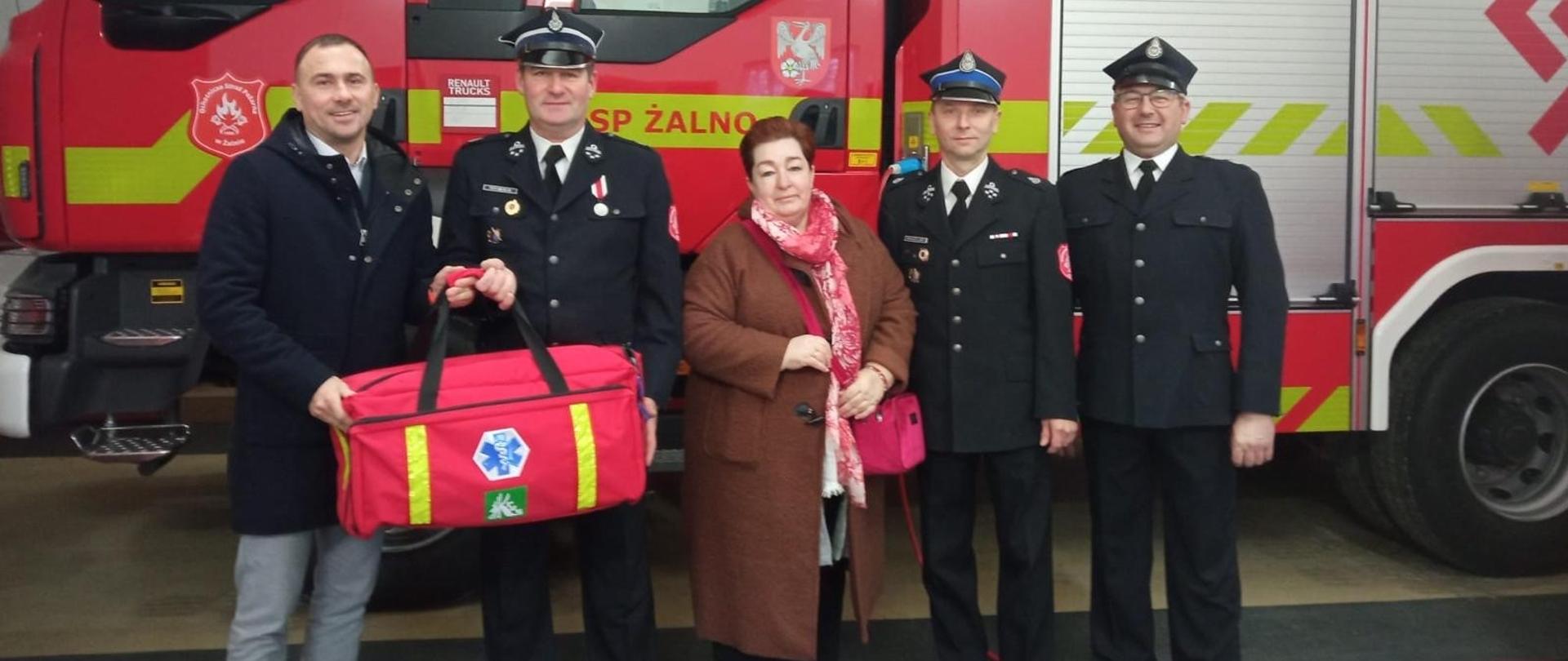 Kobieta i czterech mężczyzn, w tym trzech w mundurach galowych OSP pozują na tle wozu bojowego straży pożarnej. Stojący po lewej stronie mężczyzna w cywilnym ubraniu trzyma w rękach dużą czerwoną torbę z zestawem ratunkowym.