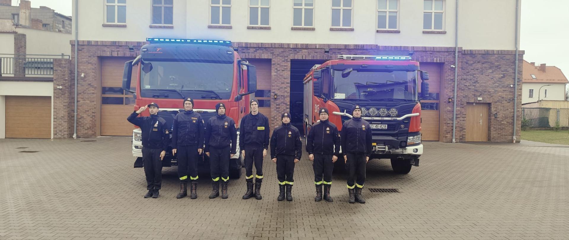 Funkcjonariusze z JRG w Pucku ustawieni w szeregu stoją na baczność. Natomiast dowódca pierwszy z lewej oddaje honor. Strażacy są ubrani w ubrania koszarowe. Na drugim planie ustawione dwa wozy bojowe.