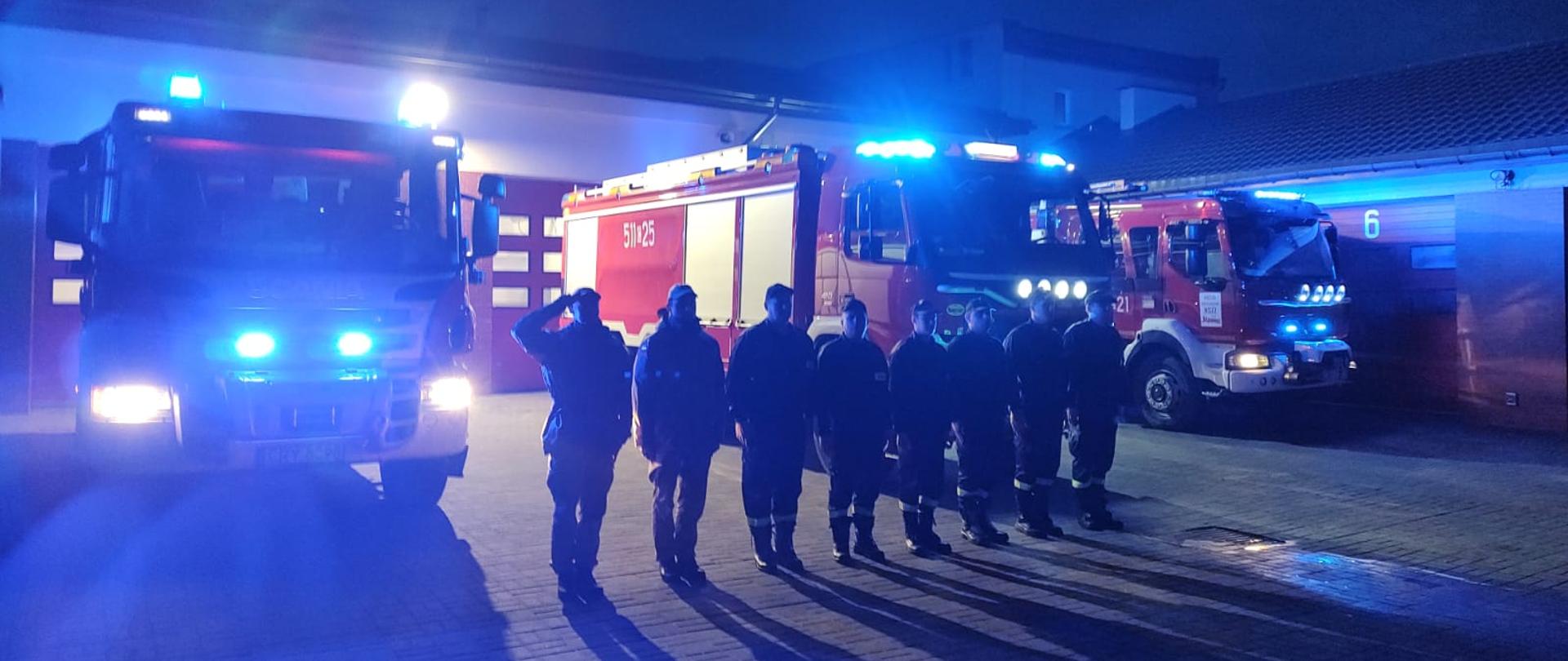 Na zdjęciu znajdują się strażacy oddający hołd zmarłemu ratownikowi medycznemu. W tle znajdują się samochody pożarnicze z włączonymi światłami alarmowymi. 