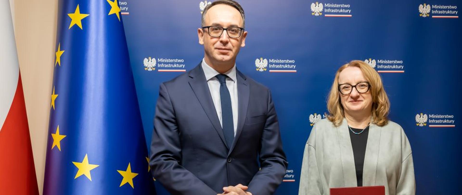 Minister Infrastruktury Dariusz Klimczak oraz nowa prezes Wód Polskich Joanna Kopczyńska stoją na granatowym tle z logiem Ministerstwa Infrastruktury. Na lewo flagi Unii Europejskiej i Polski. 