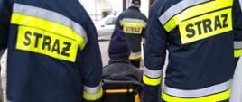 Strażacy w umundurowaniu specjalnym transportują osobę ma wózku inwalidzkim do punktu szczepień przeciw Covid-19. 