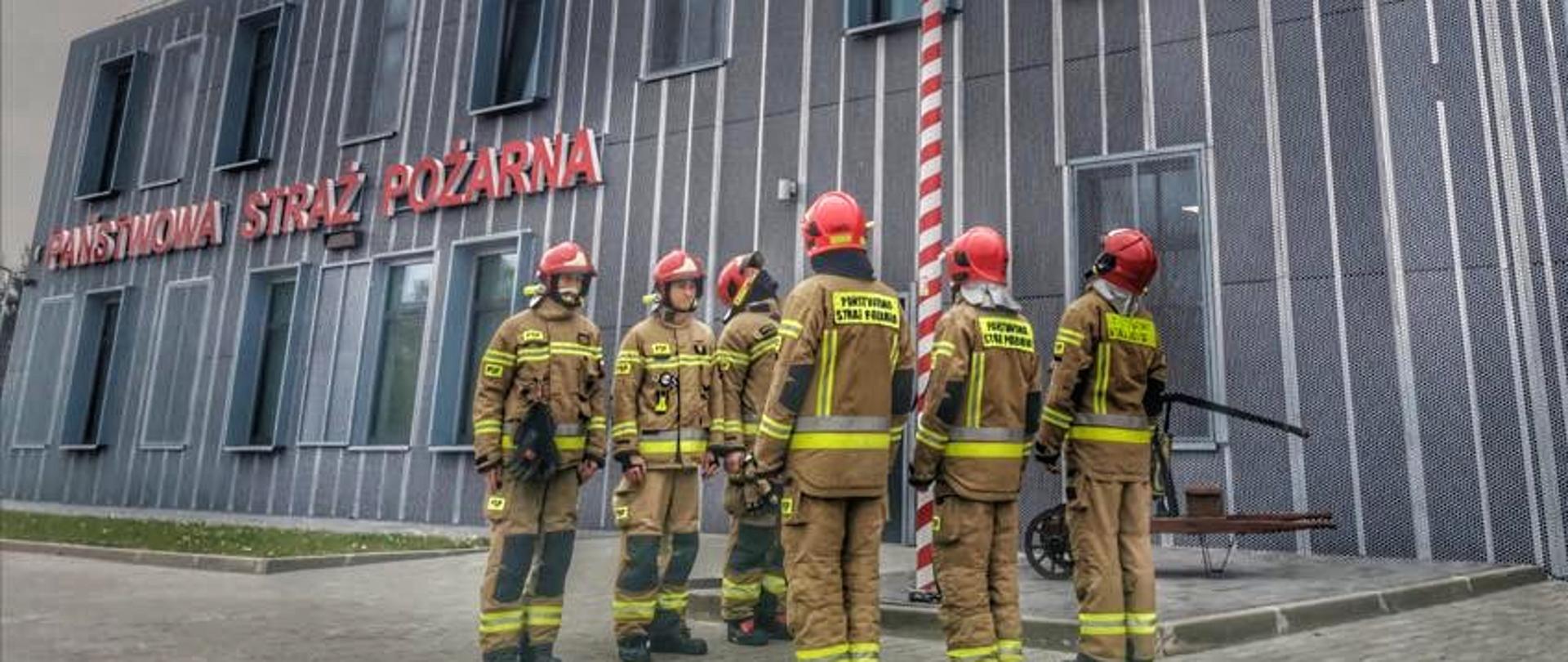 Uroczysta zmiana służby przed jednostką PSP, przy maszcie z flagą narodową.