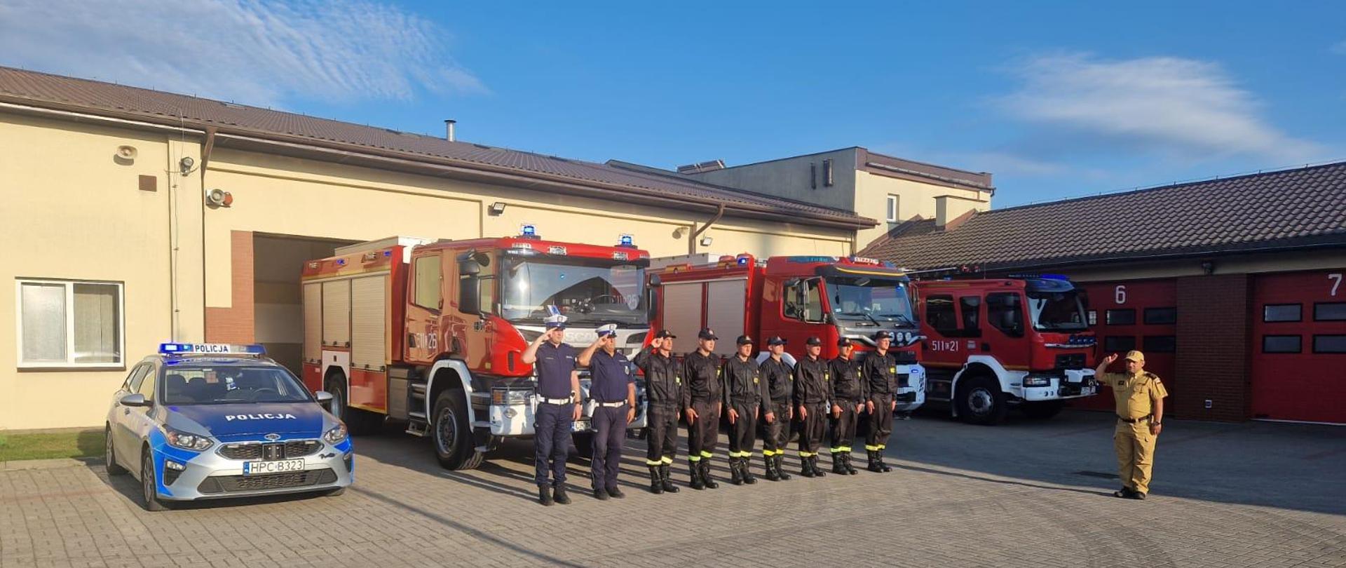 Na placu jednostki ratowniczo-gaśniczej strażacy oraz policjanci oddają hołd strażakom poległym w pożarze kamienicy w Poznaniu. Za nimi w tle wozy bojowe i budynki komendy powiatowej państwowej straży pożarnej w Rypinie.