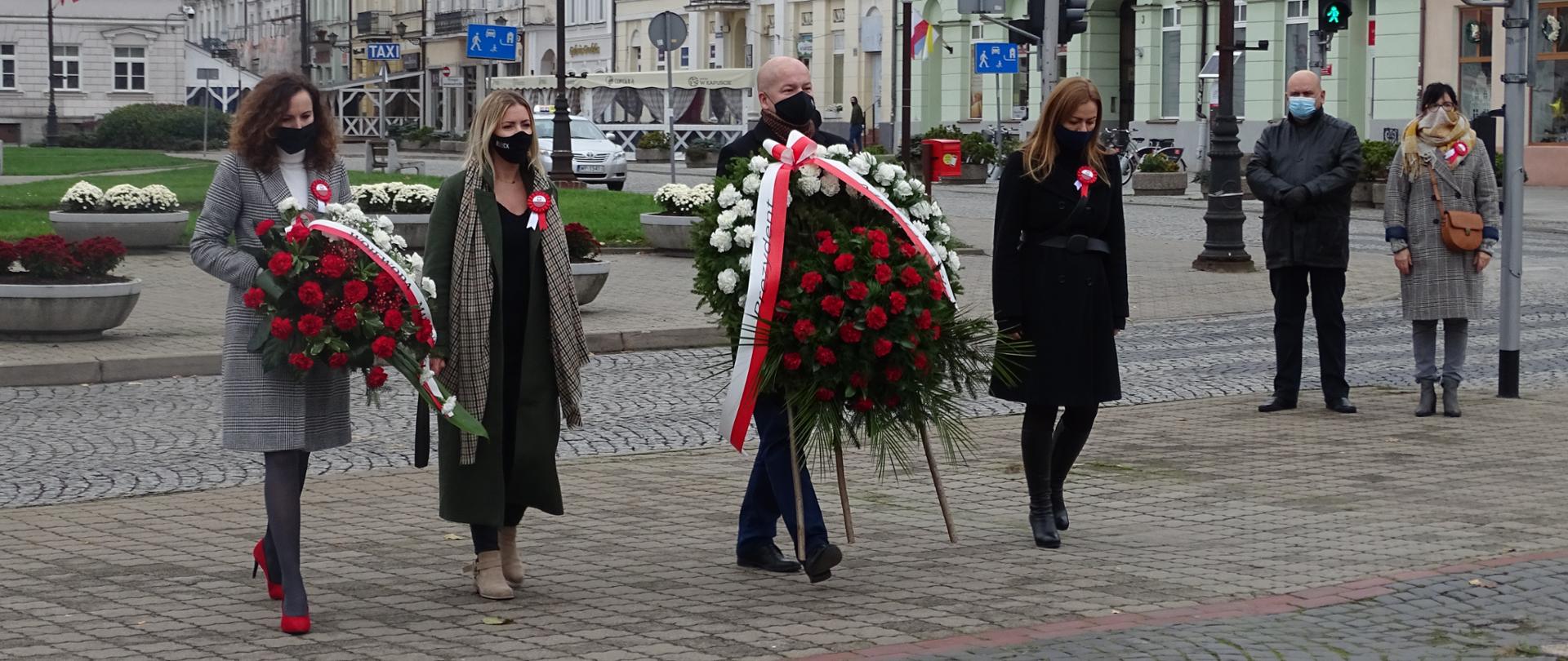 Elegancko ubrane kobiety oraz elegancko ubrany mężczyzna składają biało czerwone kwiaty. W tle widać białe budynki