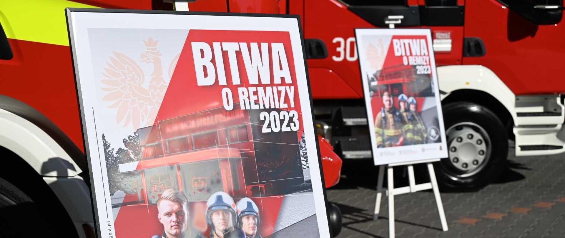 przed wozami strażackimi stoją dwa plakaty z napisem bitwa o remizy 2023 na plakacie sa trzej strażacy