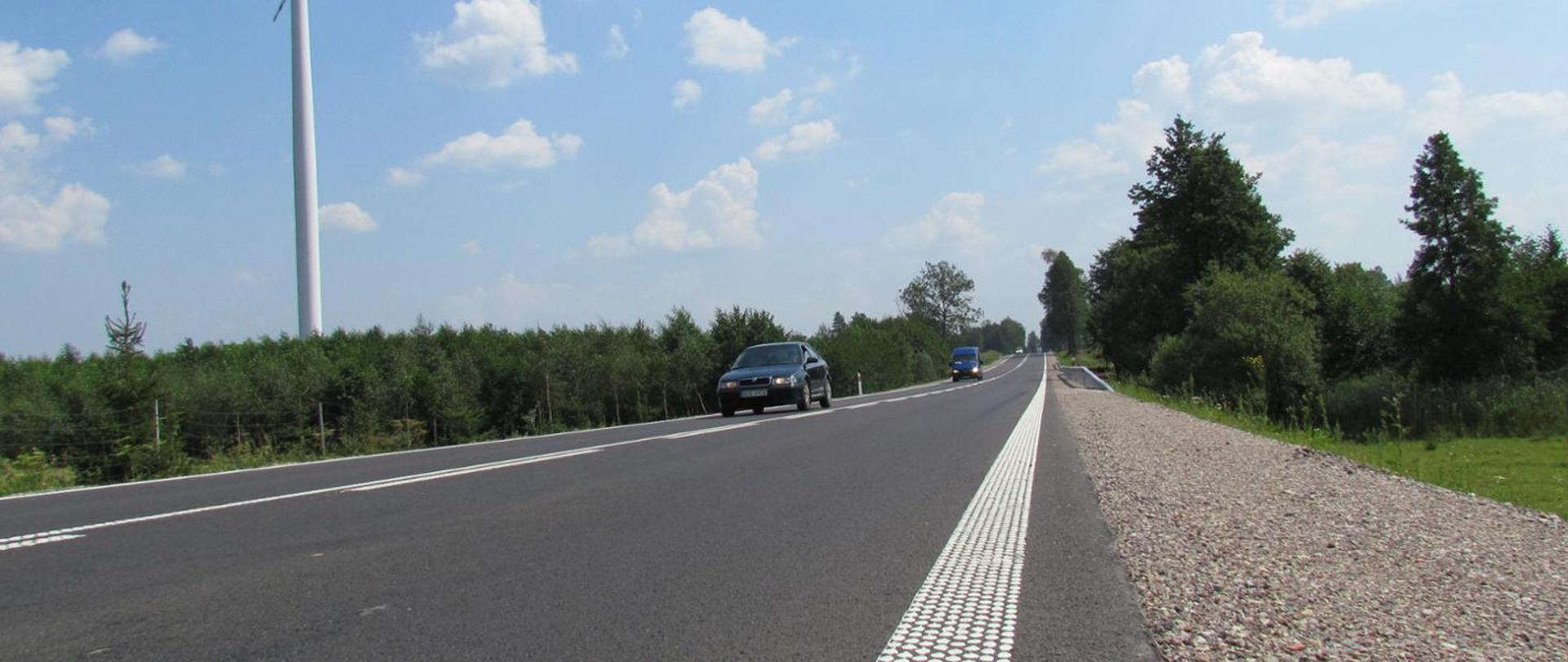 Widok z poziomu jezdni na jadące drogą pojazdy. W tle widoczne niskie drzewa i podstawa wiatraka.