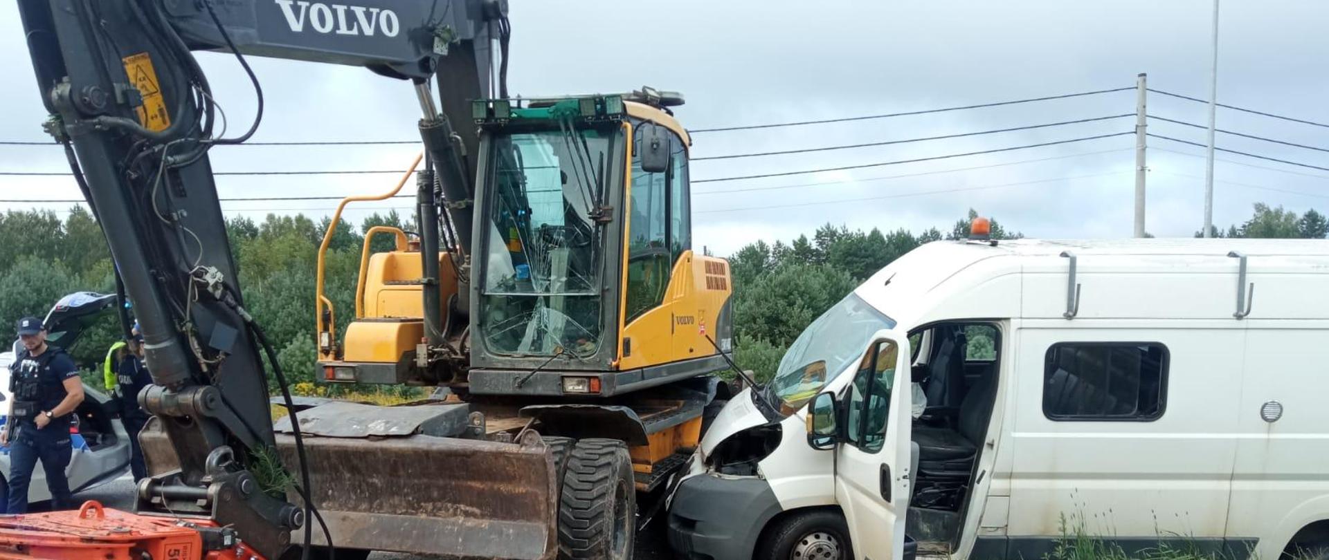 Zdjęcie przedstawia dwa uszkodzone pojazdy biorące udział w zderzeniu. Po lewej stoi koparka, a po prawej biały dostawczy bus.