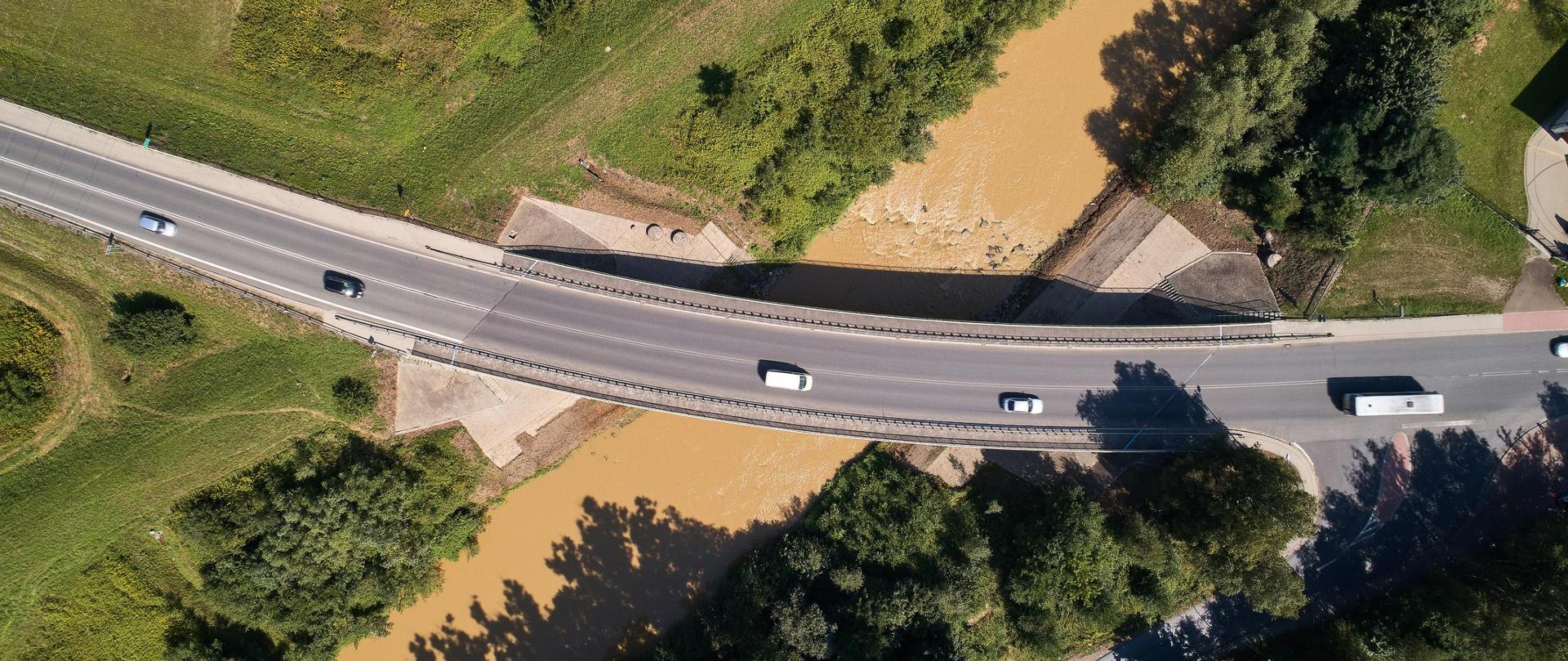 Jest lato, świeci słońce. Widok z góry na most nad rzeką Ropa w Jaśle. Most przebiega w łuku nad rzeką. Woda w rzece jest brunatna, dookoła zielone drzewa, trawniki. Na moście jadą auta.