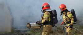 Zdjęcie przedstawia dwóch strażaków w trakcie ćwiczeń. Niosą linię gaśniczą zakończoną srebrną wytwornicą. Umundurowani w mundury koloru piaskowego z założonymi maskami sprzętu ochrony dróg oddechowych z butlami powietrznymi. Mają na sobie czerwone hełmy. W tle widoczny dym sceniczny.
