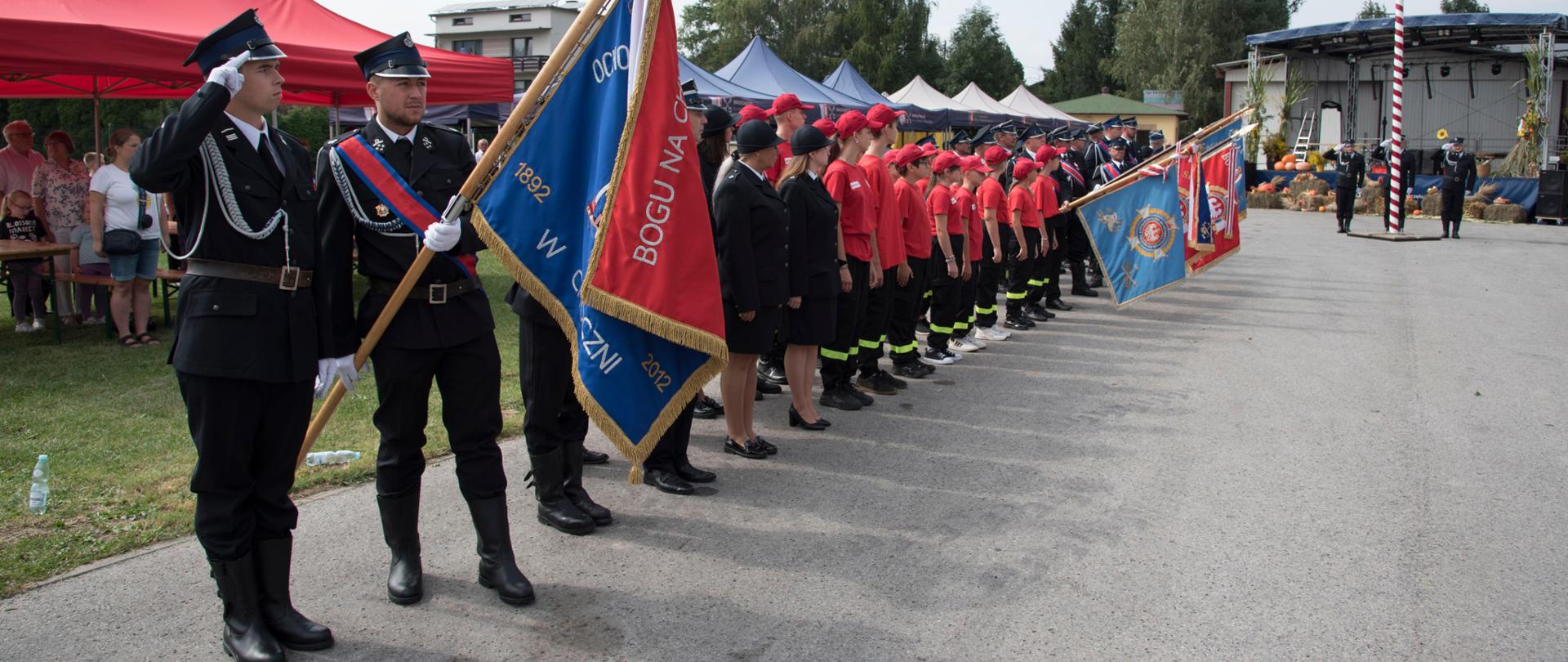 Po lewej pododział strażaków w mundurach wyjściowych. Poczet sztandarowy salutuje. W tle po prawej widoczny maszt i salutujący poczet flagowy