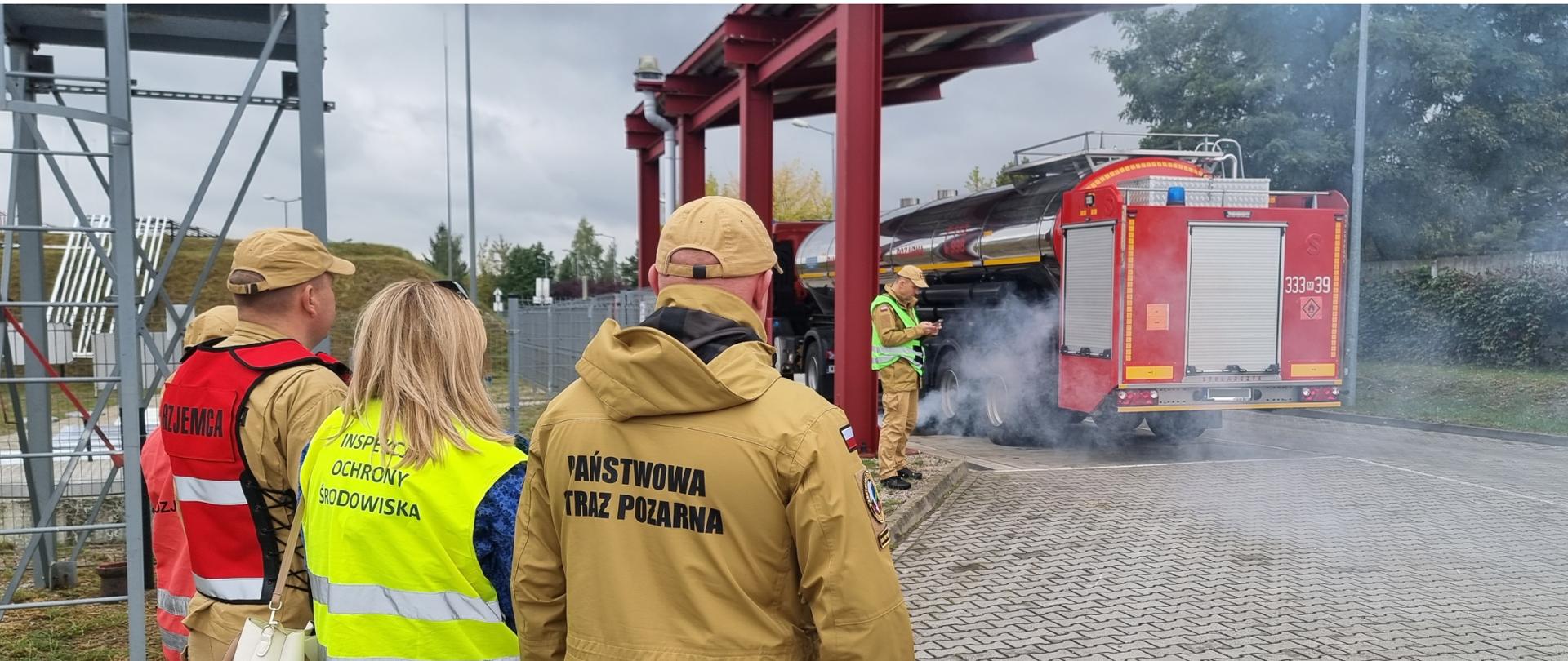 Inspektor Wojewódzkiego Inspektoratu Ochrony Środowiska w Warszawie wraz z funkcjonariuszami Państwowej Straży Pożarnej stoi kilkanaście metrów od wozu PSP i monitoruje akcję gaśniczą.