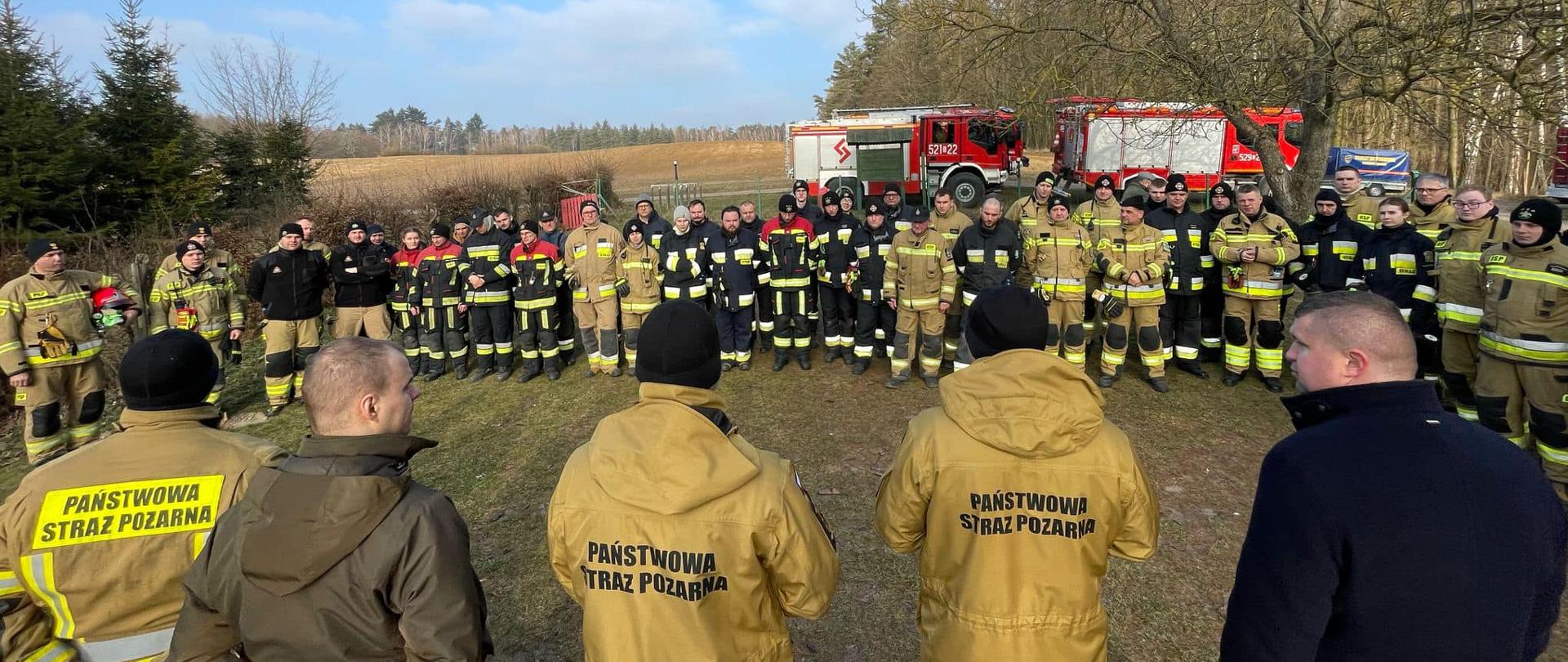 Odprawa z prowadzącymi ćwiczenia, strażacy ustawieni w szeregu, w tle las i samochody strażackie