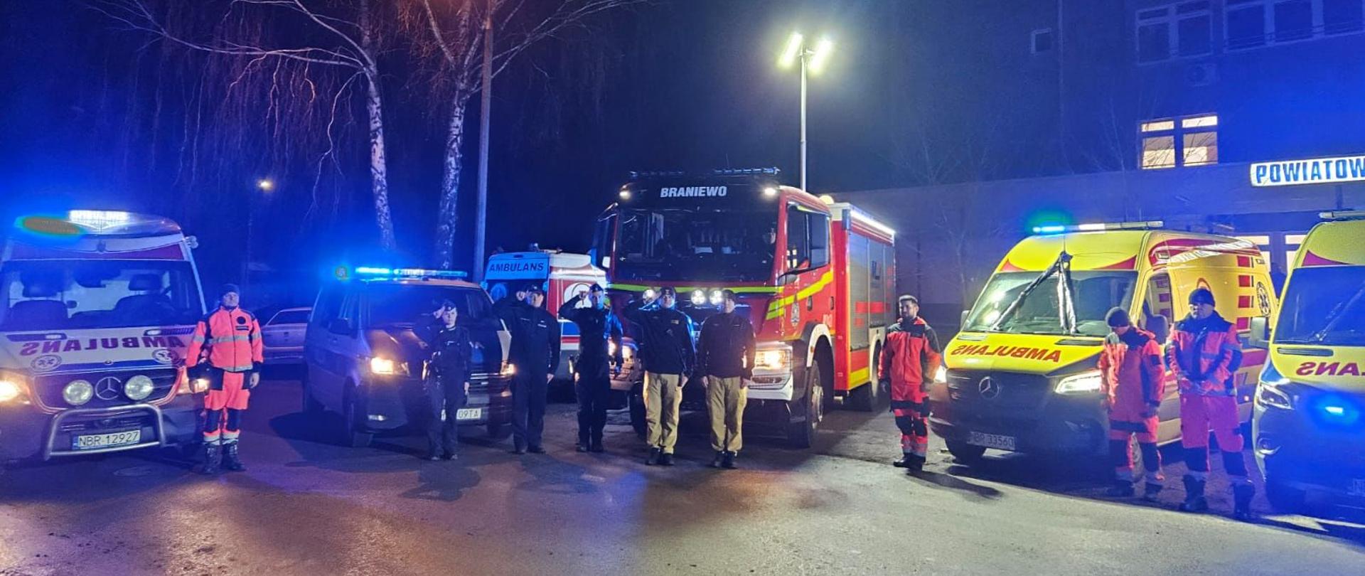 Samochody strażackie, karetki z włączonymi światłami i ratownikami przy samochodach.