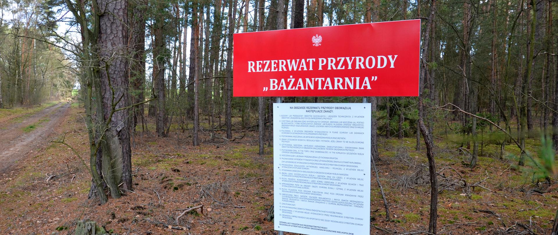 Czerwona tablica z napisem Rezerwat przyrody Bażantarnia w tle drzewa liściaste.