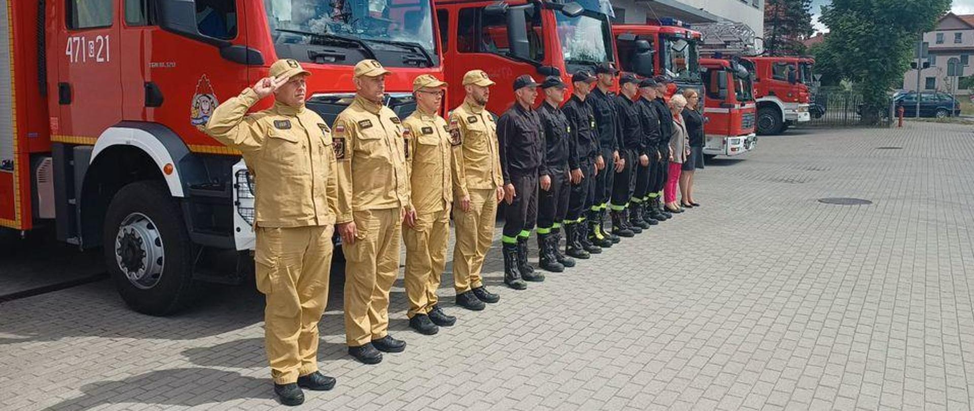 Strażacy i pracownicy służby cywilnej stoją na baczność w rzędzie przed budynkiem komendy. Za nimi widoczne samochody ratowniczo - gaśnicze. Pierwszy z lewej strażak salutuje.