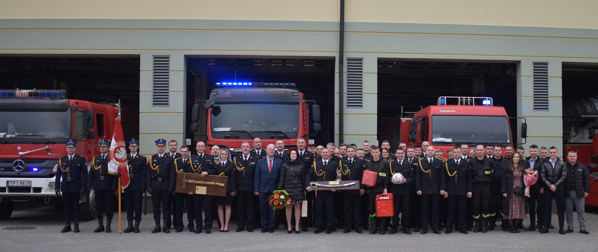 Zdjęcie przedstawia uczestników apelu z okazji przejścia na zaopatrzenie emerytalne zastępcy dowódcy JRG. Strażacy PSP występują w umundurowaniu wyjściowym. W tle widać samochody pożarnicze. 