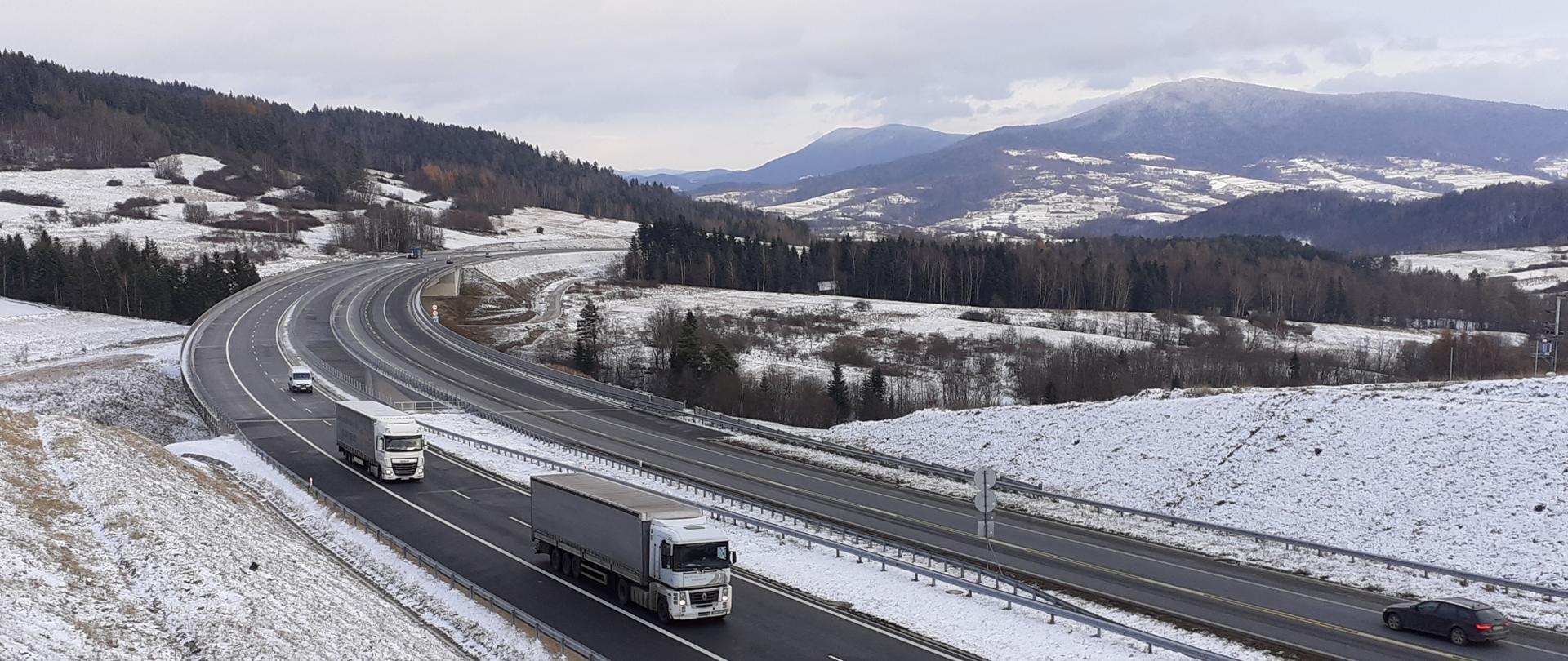 Widok na dwujezdniowy odcinek S7 z poruszającymi się pojazdami w obu kierunkach z przepięknym krajobrazem w tle. Teren pokryty pierwszym śniegiem. 