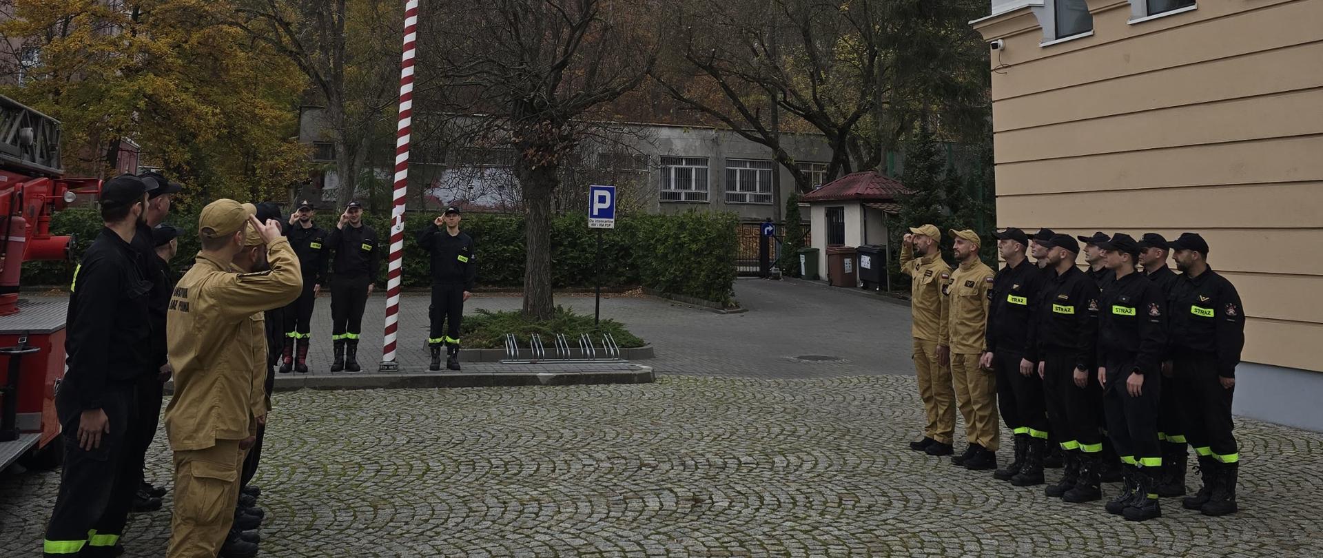 Strażacy stoją w szeregu podczas zmiany służby. W tle maszt z flagą Polski.