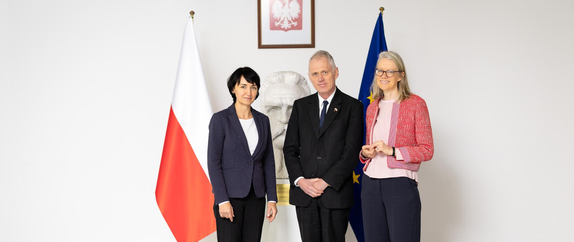 Wiceminister Henryka Mościcka-Dendys stoi z dwiema osobami na tle dwóch flag. 