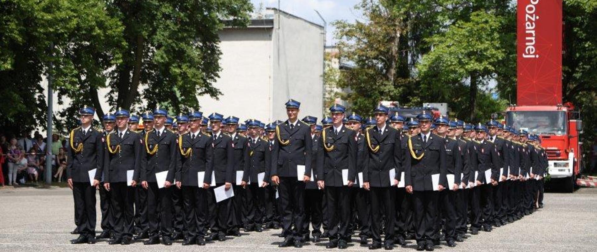 Funkcjonariusze Państwowej Straży pożarnej w mundurach wyjściowych w kolumnach czwórkowych na placu centralnej szkoły państwowej straży pożarnej w częstochowie.