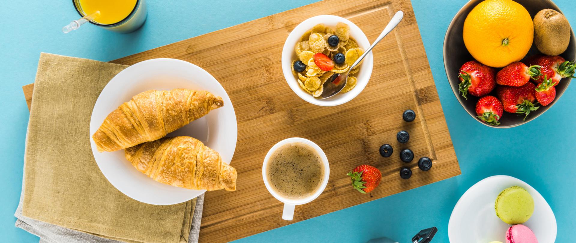 Na zdjęciu widoczne jest śniadanie. Na desce do krojenia leżą rogale, płatki, owoce oraz stoi kawa. Ponadto obok jest kawiarka, miska z owocami, makaroniki i sok pomarańczowy. Tło jest niebieskie.