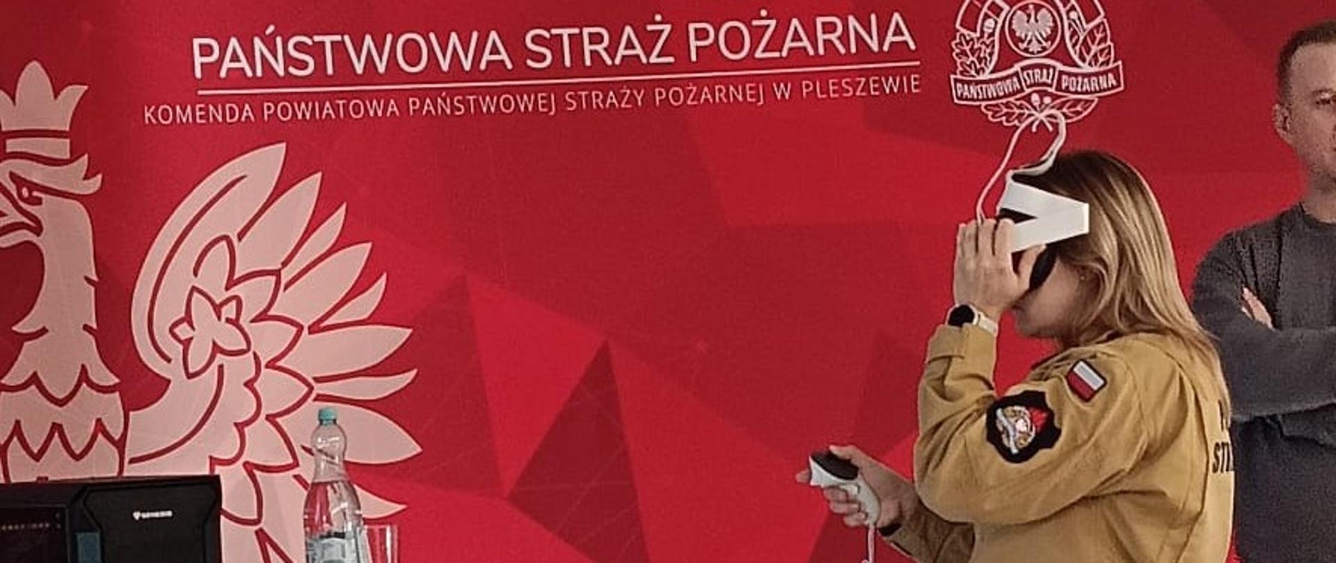 Kobieta w żółtym mundurze Państwowej Straży Pożarnej stoi z goglami VR na głowie, obok stoi mężczyzna w ubraniu cywilnym, za nimi widać czerwony baner z napisem Państwowa Straż Pożarna