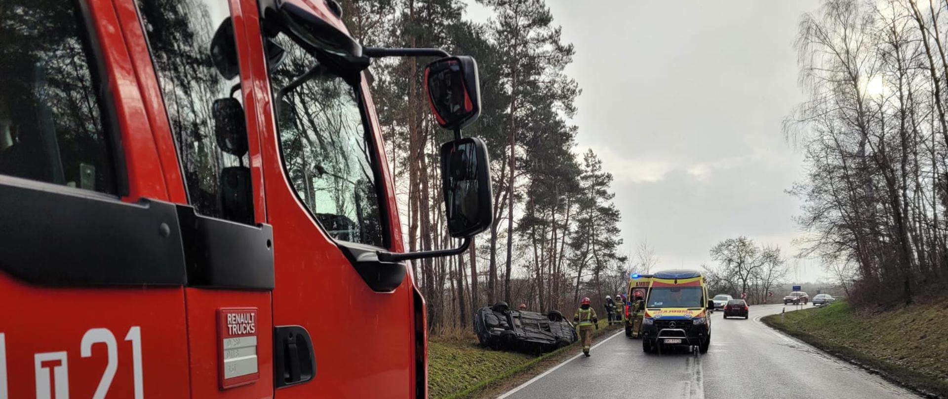 Samochód Pożarniczy, w tle dwa ambulanse, samochód biorący udział w zdarzeniu a także strażaków pracujących na miejscu zdarzenia.