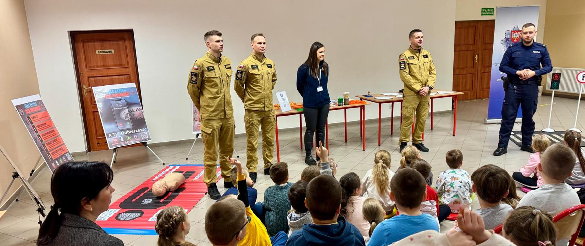 Na zdjęciu widzimy spotkanie edukacyjne skierowane do dzieci, które siedzą na krzesełkach i uważnie słuchają prelegentów. W centrum stoją cztery osoby – trzech mężczyzn w mundurach straży pożarnej, jedna osoba z identyfikatorem, oraz policjant. W tle widoczne są plansze edukacyjne, w tym manekin do nauki pierwszej pomocy oraz znaki drogowe, co sugeruje, że spotkanie dotyczy bezpieczeństwa i udzielania pierwszej pomocy. Sala jest przestronna, a atmosfera wydaje się być przyjazna i edukacyjna.