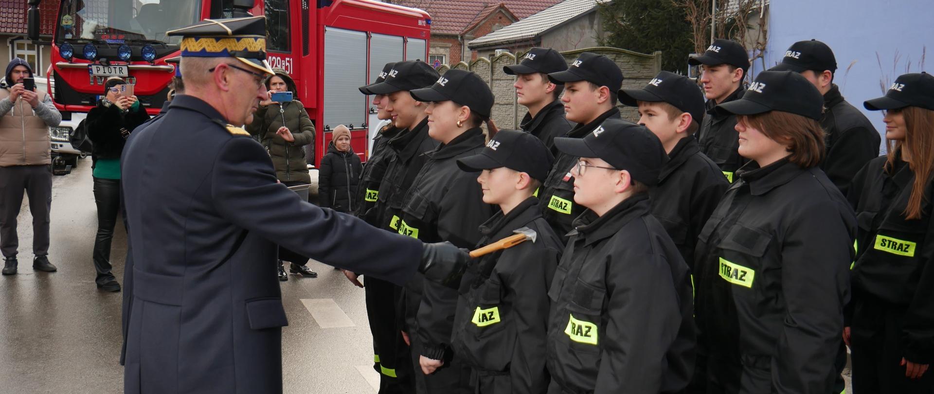 Lubuski komendant wojewódzki w ciemnym płaszczu z dystynkcjami trzyma toporek ceremonialny i oficjalnie dotyka nim ramienia młodej osoby w szeregu. Grupa młodych ludzi ubranych w czarne uniformy z napisem "STRAŻ" stoi w szeregu na ulicy, obok znajduje się czerwony wóz strażacki z napisem „OSP RUSINÓW”. W tle stoją mieszkańcy, w tym osoby robiące zdjęcia.