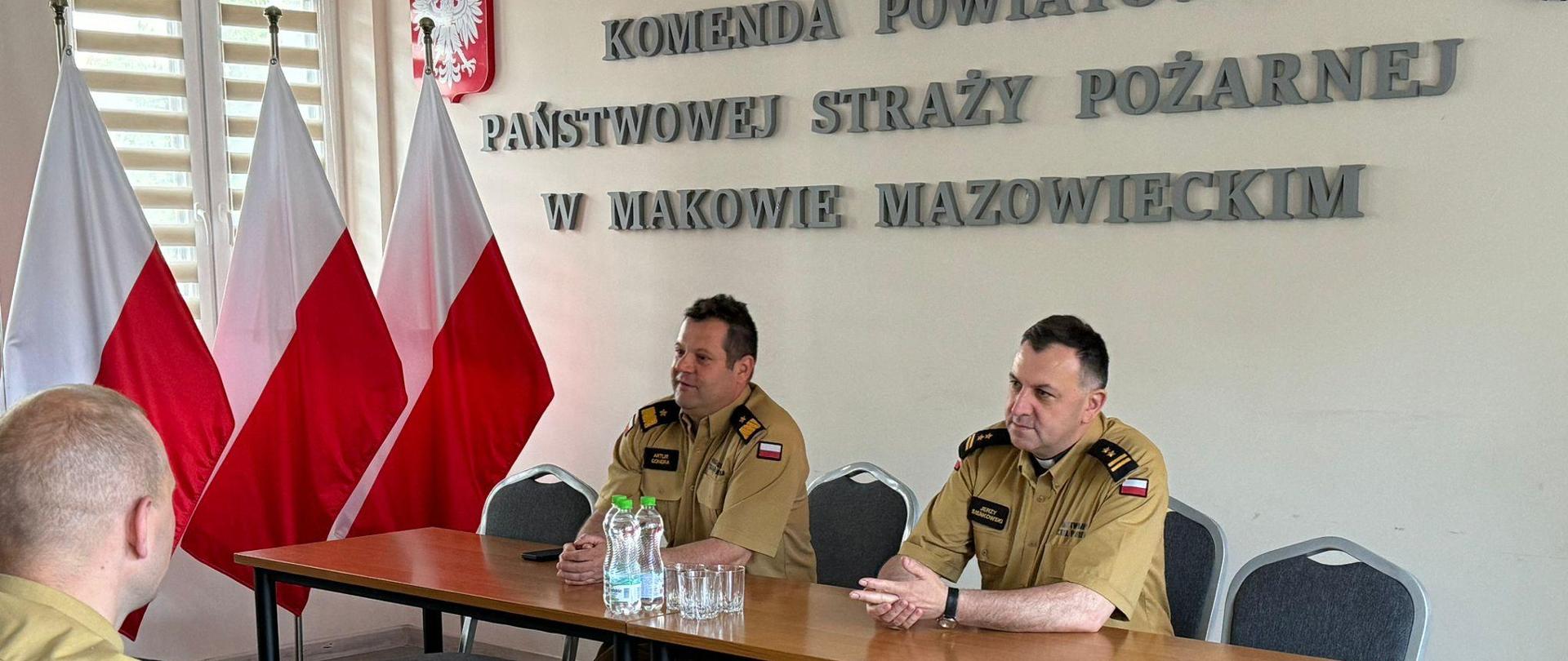 Mazowiecki Komendant Wojewódzki oraz Kapelan Mazowieckich Strażaków siedzą na świetlicy przy stole. Przed nimi siedzą w ławkach strażacy.