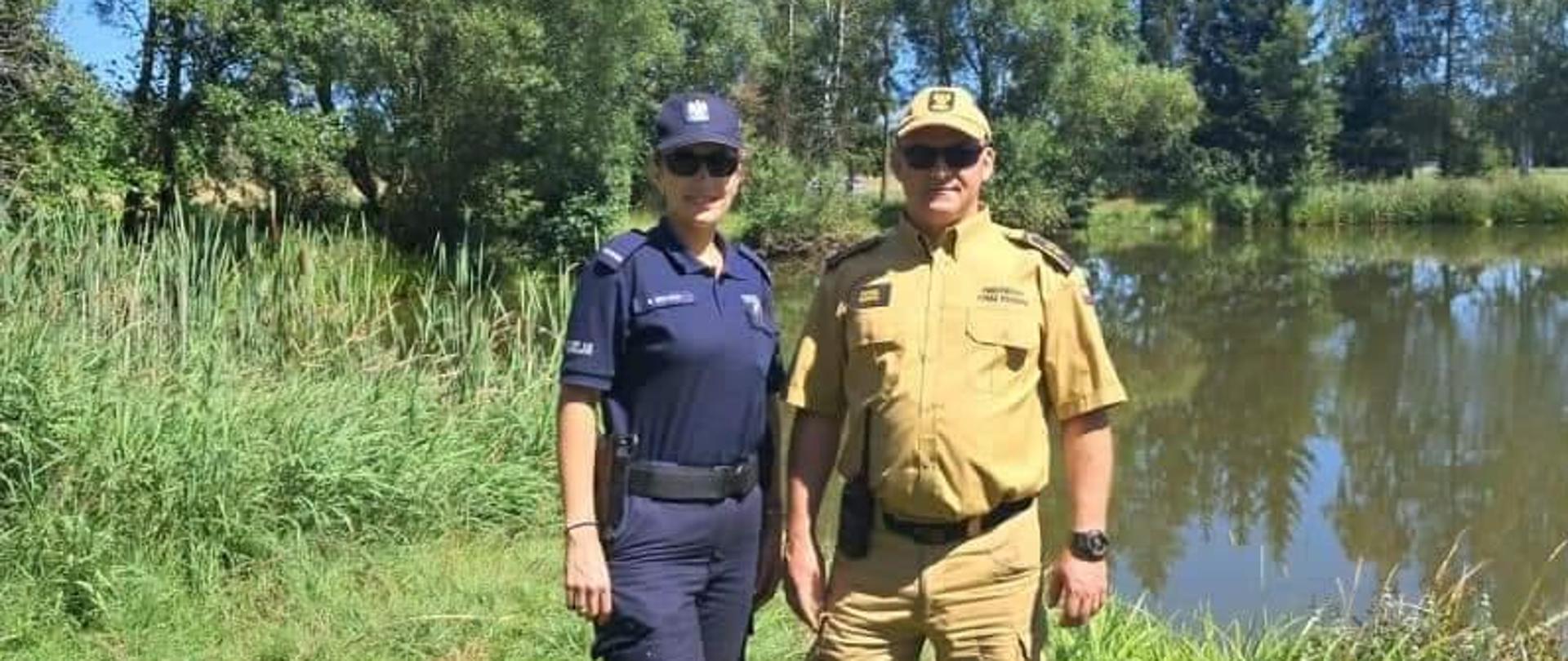 Funkcjonariusz straży pożarnej oraz funkcjonariuszka policji na planie głównym w tle zbiornik wodny. Zdjęcie wykonane w terenie podczas wspólnych patroli zbiorników wodnych.