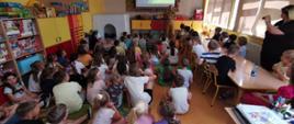 Strażak w sali lekcjyjnej opowiada dzieciom, włączona prezentacja na projektorze multimedialnym. Dzieci siedzą przy stolikach i na podłodze. Ściany kolorze żółtym, gazetka na ścianie lewej. Szafeczki z kolorowymi frontami.