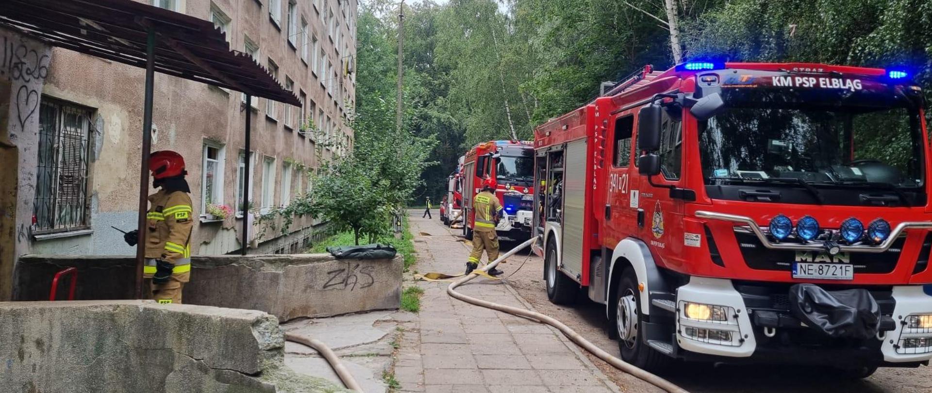 Po lewej stronie znajduje się budynek wielorodzinny, czteropiętrowy, obok znajdują się sylwetki strażaków w piaskowych ubraniach specjalnych i czerwonych hełmach. Po prawej stronie, wzdłuż ulicy stoją zaparkowane, trzy czerwone pojazdy pożarnicze.