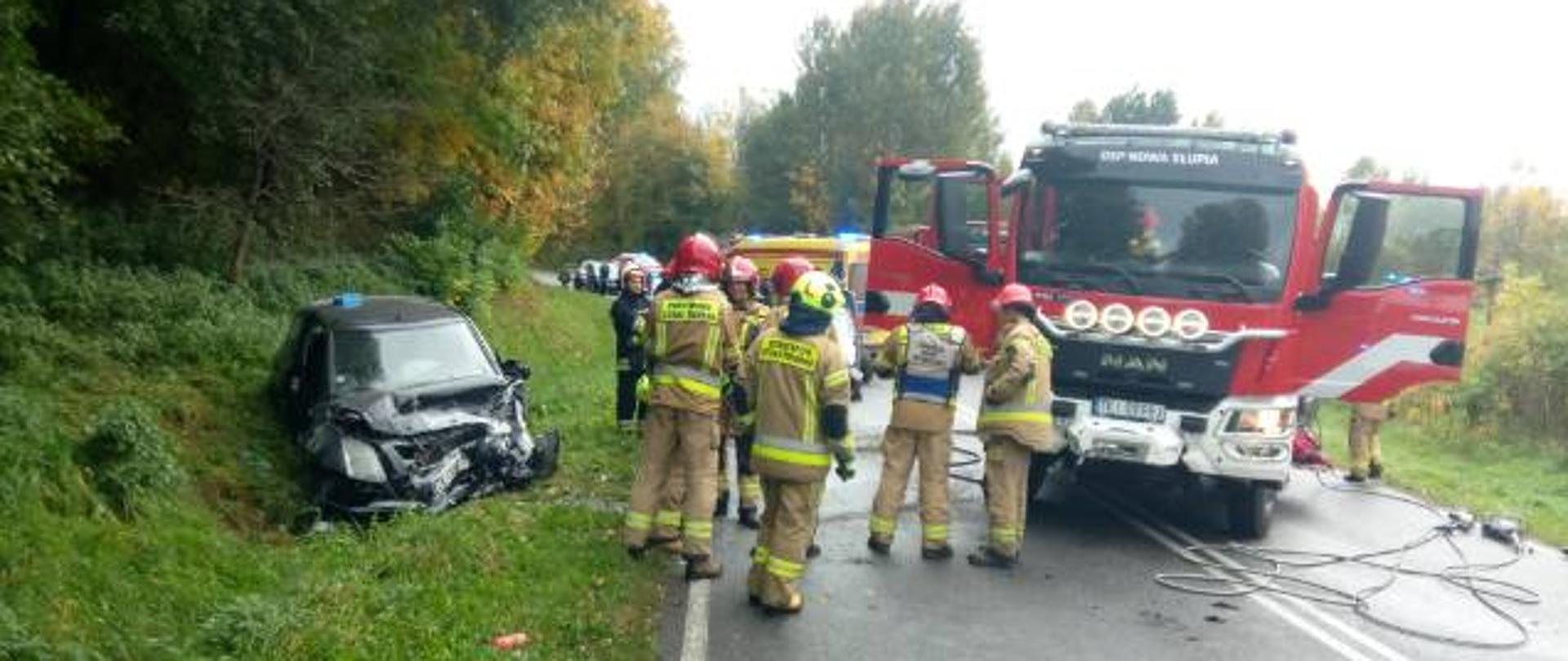 Zdjęcie przedstawia drogę wojewódzką, na której stoją samochody służb ratunkowych – po prawej stronie, w centralnym punkcie grupa ratowników a z lewej widać znajdujący się w przydrożnym rowie jeden z uszkodzonych samochodów.