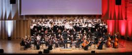 Scena w filharmonii - chór i orkiestra szkolna z dyrygentem. W tle ekran z tekstem pieśni