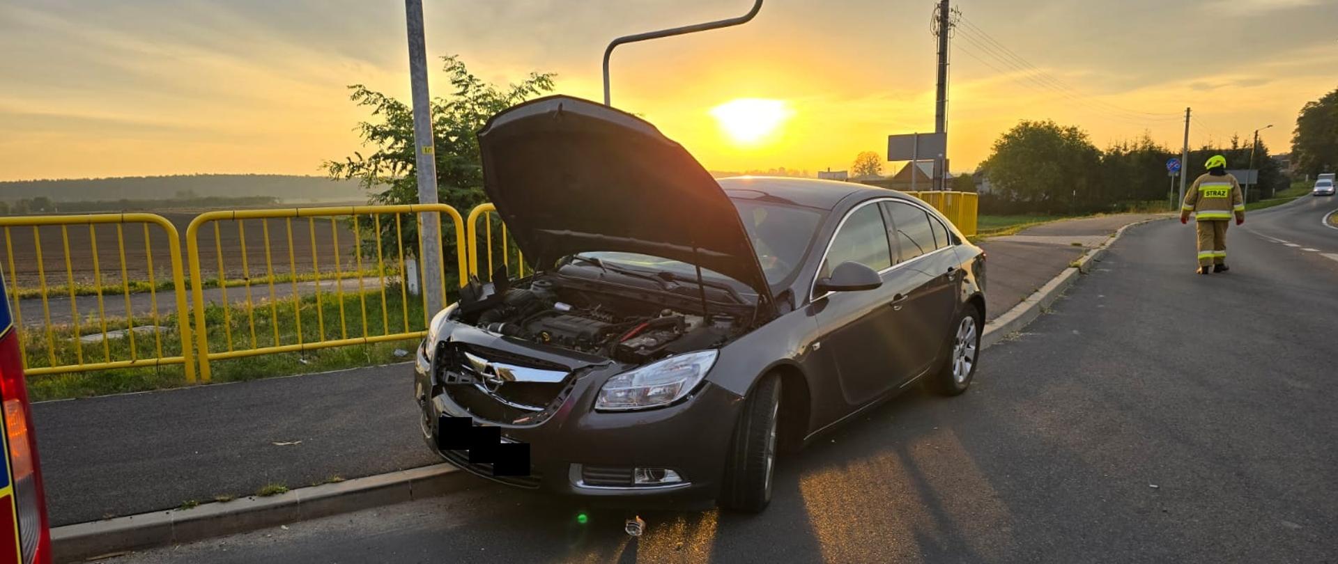 Uszkodzony czarny samochód na skrzyżowaniu, za nim strażak na drodze