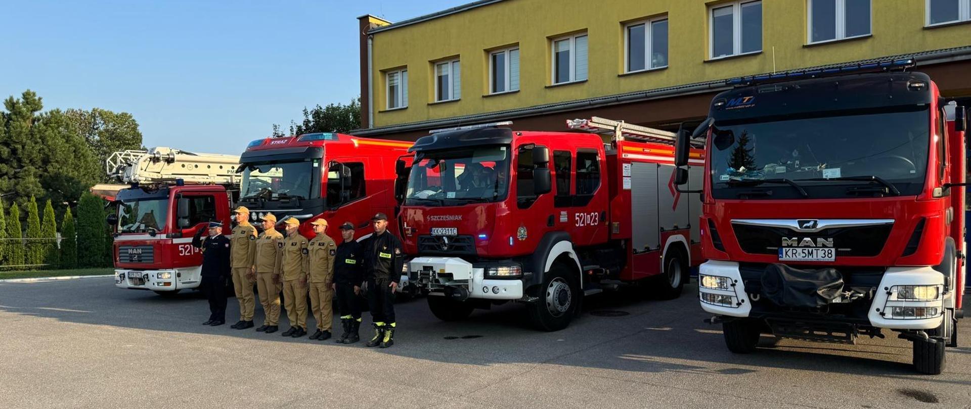 Strażacy stoją w szeregu na tle samochodów strażackich