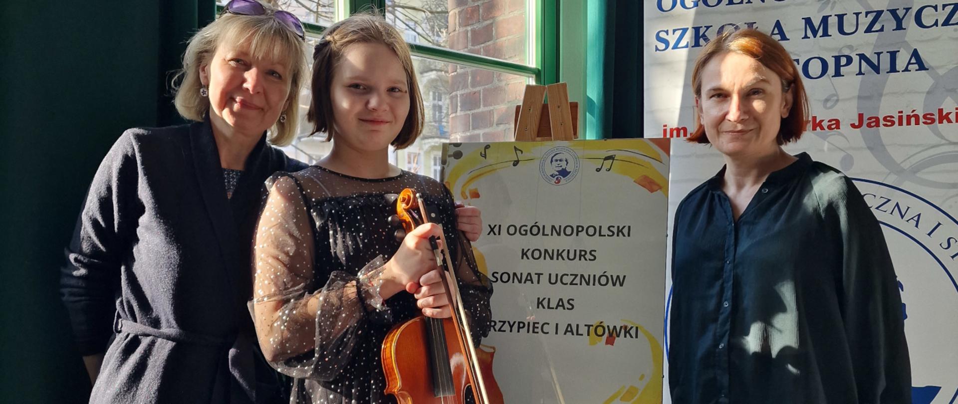 Na zdjęciu uczennica Antonina Wojciul. Po obu stronach uczennicy stoją nauczycielki Olga Borowska i Magdalena Borowska. Uczennica trzyma skrzypce. Na sztaludze wisi plakat z informacją o konkursie.