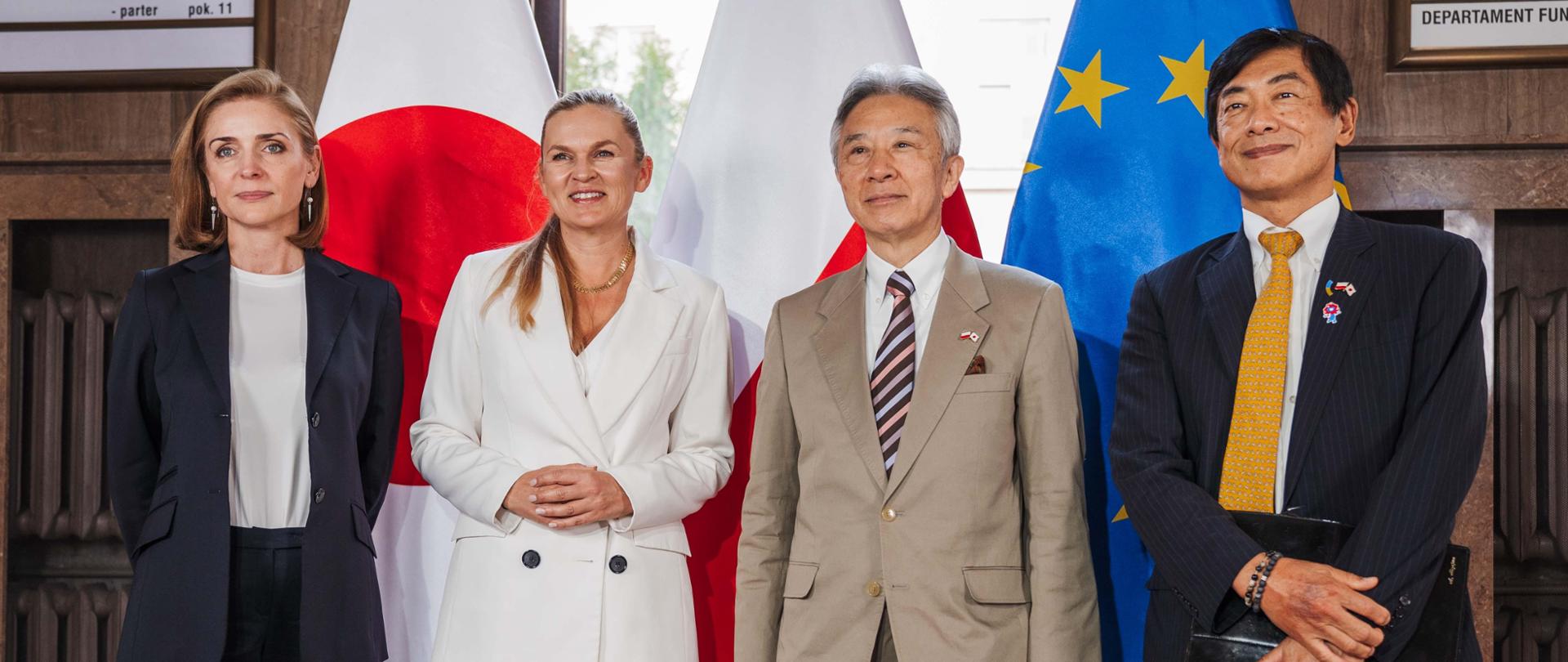 Na tle flag Polski i Japonii stoi minister Barbara Nowacka, wiceminister Joanna Mucha i japoński Minister Edukacji, Kultury, Sportu, Nauki i Technologii oraz mężczyzna w garniturze. Pozują do zdjęcia.