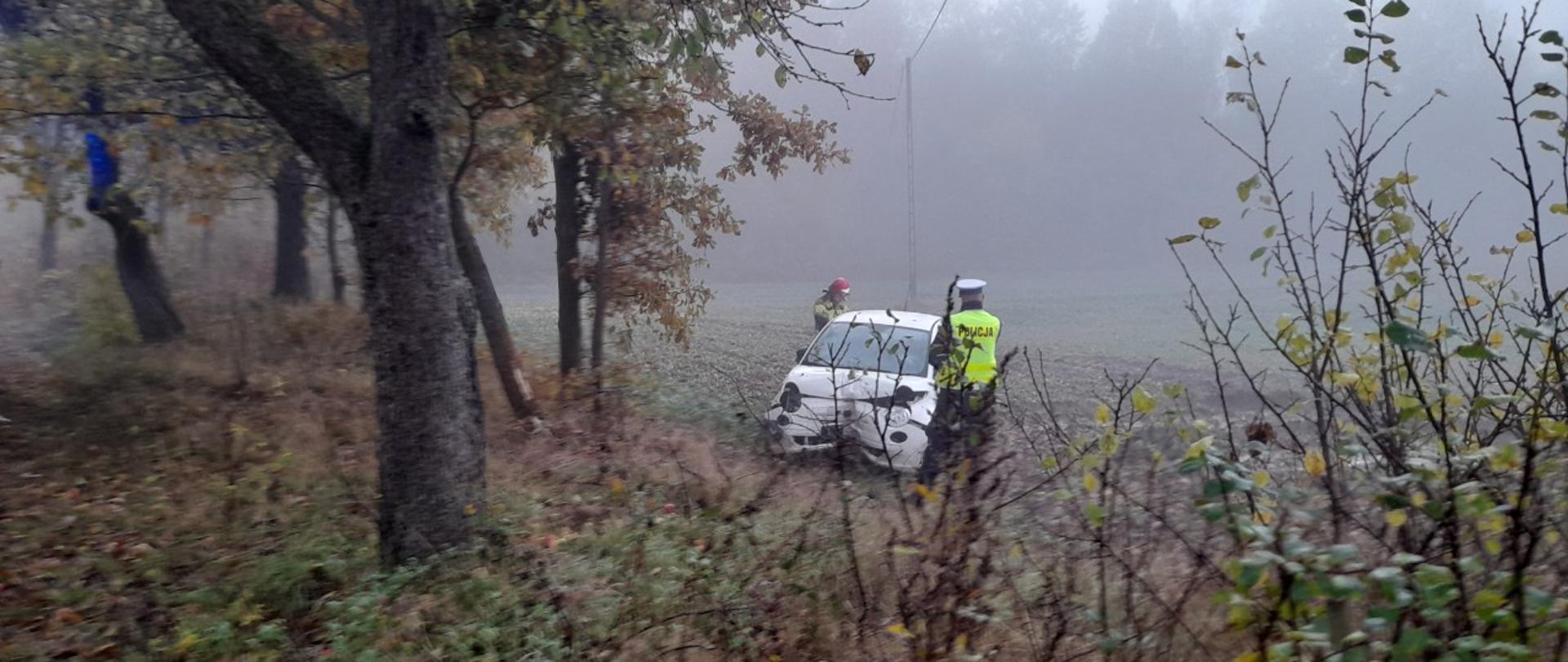 Uszkodzony pojazd stoi na polu, przy pojeździe strażak i policjant