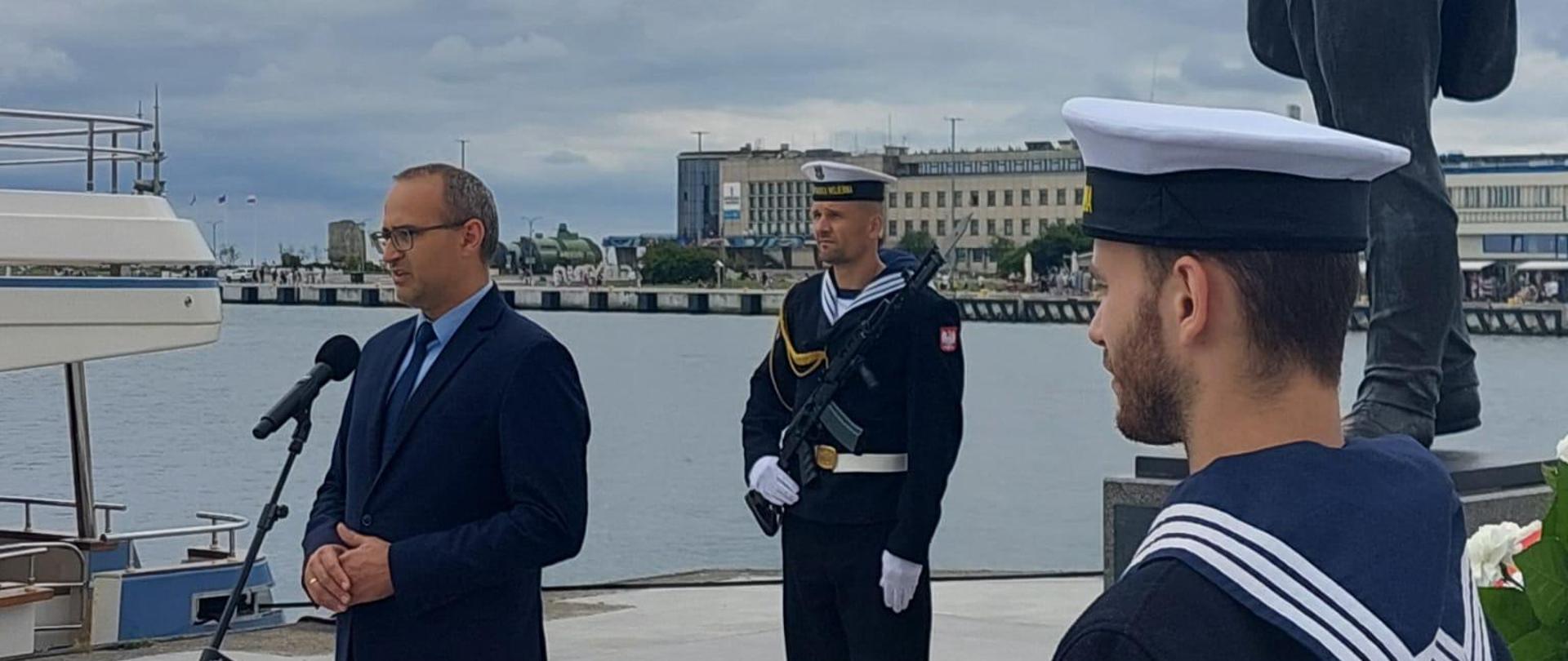 mężczyzna stoi przed mikrofonem i przemawia, w tle kanał portowy i marynarz stojący przy pomniku budowniczego portu