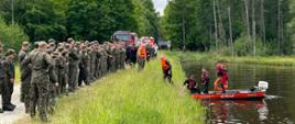 Na zdjęciu widzimy strażaków ubranych w skafandry wypornościowe którzy wodują łódź na zbiorniku wodnym. Obok niego znajduje się torba medyczna. Na brzegu, w dwuszeregu zgromadzeni są żołnierze, którzy obserwują działania strażaków. W tle widoczny jest las.