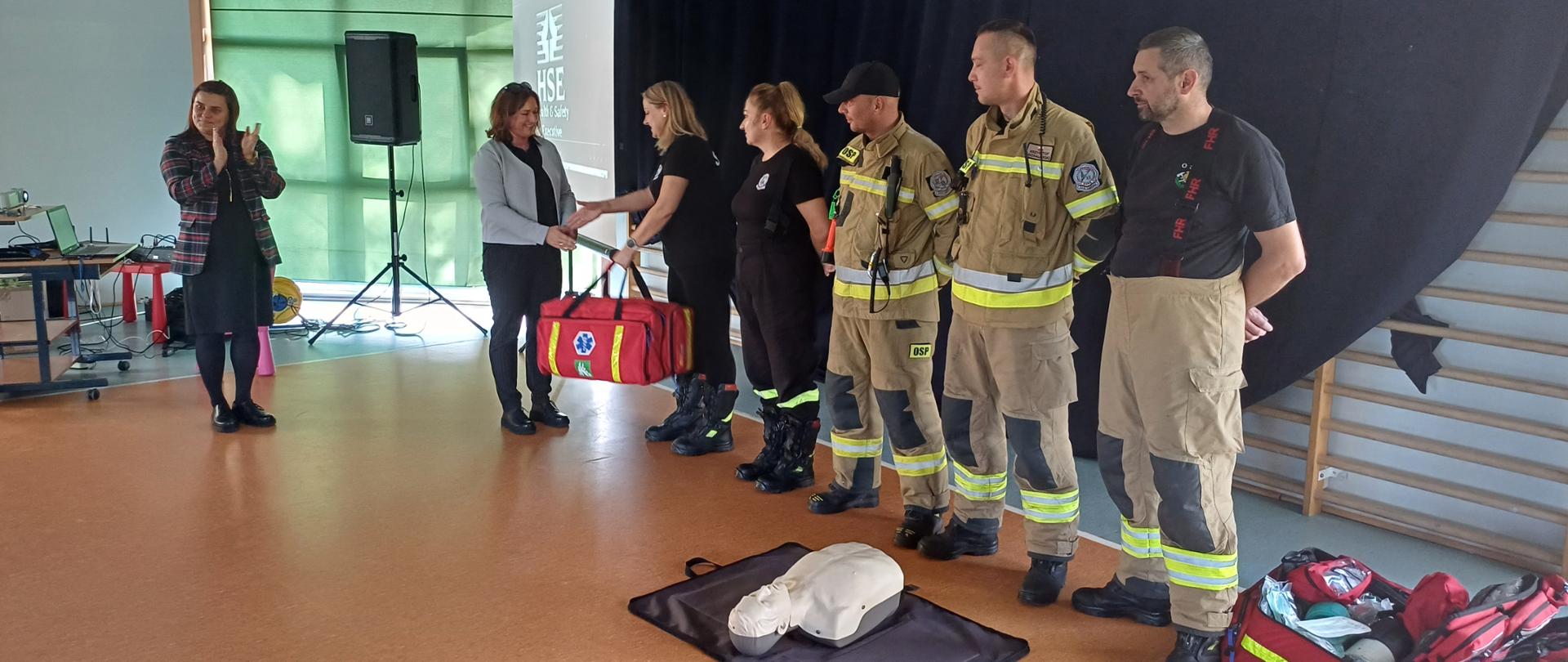 W sali gimnastycznej stojąca z lewej kobieta bije brawo, stojąca po jej prawej stronie kobieta wręcza strażaczce torbę z zestawem ratownictwa przedlekarskiego. Obok nich stoi jeszcze jedna strażaczka i trzech strażaków. Przed nimi na podłodze leży fantom szkoleniowy.