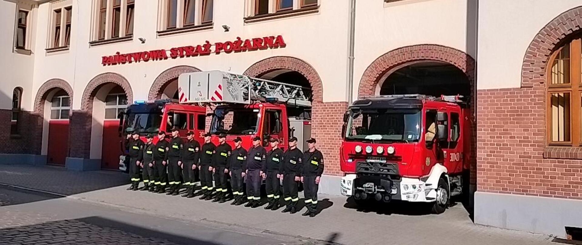 Zbiórka przed budynkiem JRG1. Strażacy stoją w szeregu w czarnych ubraniach koszarowych, za nimi przody trzech pojazdów pożarniczych stojących w bramach